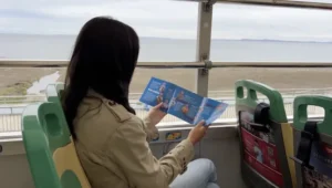 Mujer en un bus turístico mira un folleto de paradas mientras observa el paisaje costero desde la ventana.