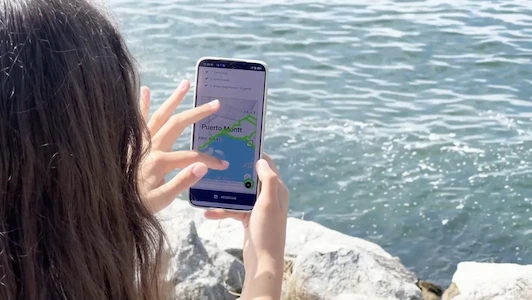 Mujer sostiene un smartphone mostrando la App Turistik Chile con imagen de bus rojo, frente a un cuerpo de agua.