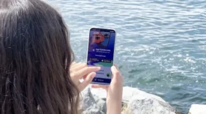 Mujer sostiene un smartphone mostrando la App Turistik Chile con imagen de bus rojo, frente a un cuerpo de agua.