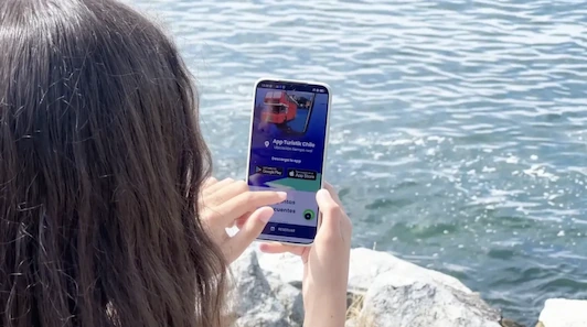 Mujer sostiene un smartphone mostrando la App Turistik Chile con imagen de bus rojo, frente a un cuerpo de agua.