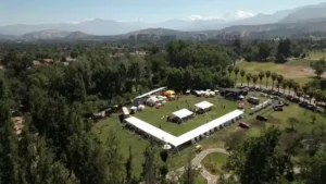 Vista aérea de un evento al aire libre con carpas blancas en forma de U en un parque rodeado de árboles y montañas.
