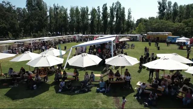 Vista aérea de un evento masivo al aire libre con sombrillas blancas, mesas y público en un parque arbolado.
