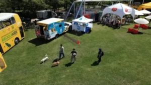 Vista aérea de un evento ParquePet y ReciclaKids con bus amarillo, unidad de veterinaria móvil y personas paseando perros sobre el césped.