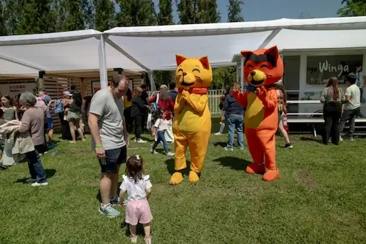 Dos mascotas de gatos naranja y amarillo interactúan con un hombre y una niña en un evento al aire libre bajo carpas blancas.