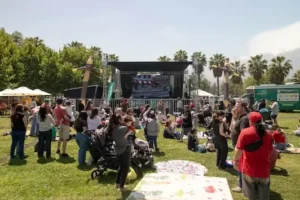 Familias con niños y carritos disfrutan de un festival al aire libre frente a un escenario con pantalla grande y muñecos inflables en un parque.