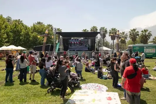 Familias con niños y carritos disfrutan de un festival al aire libre frente a un escenario con pantalla grande y muñecos inflables en un parque.