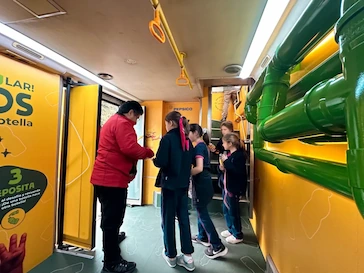 Niños y una adulta interactúan en el interior del bus ReciclaKids de TEM Turistik con paneles educativos y tuberías verdes.