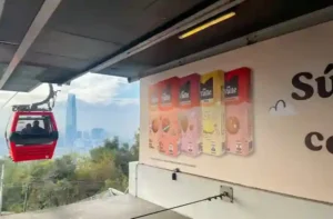 Cabina roja de teleférico en movimiento con la ciudad y el horizonte de fondo junto a un letrero publicitario en TEM Turistik.