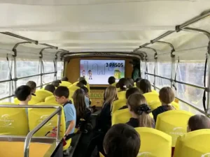 Niños sentados en el interior de un autobús adaptado viendo una presentación educativa en pantalla durante una actividad de TEM Turistik.