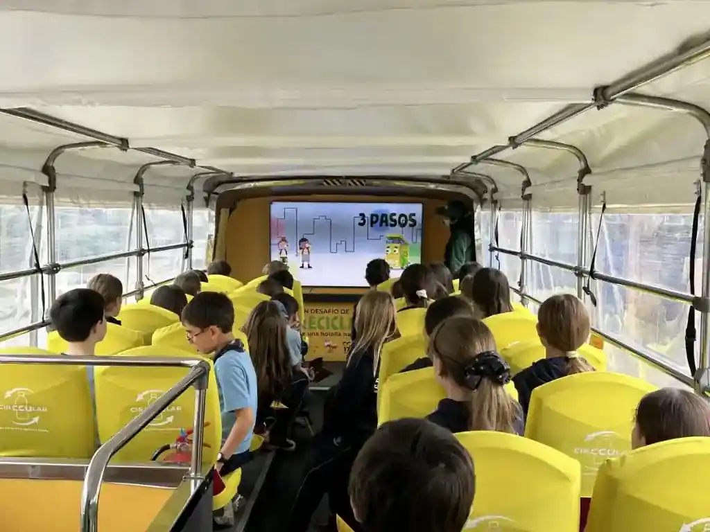 Niños sentados en el interior de un autobús adaptado viendo una presentación educativa en pantalla durante una actividad de TEM Turistik.