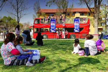 Familias sentadas en el césped disfrutando de un espectáculo artístico en vivo sobre un autobús rojo de dos pisos en TEM Turistik.