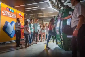Pasajeros subiendo a una cabina verde de teleférico en la estación de TEM Turistik junto a personal de apoyo y paneles publicitarios.