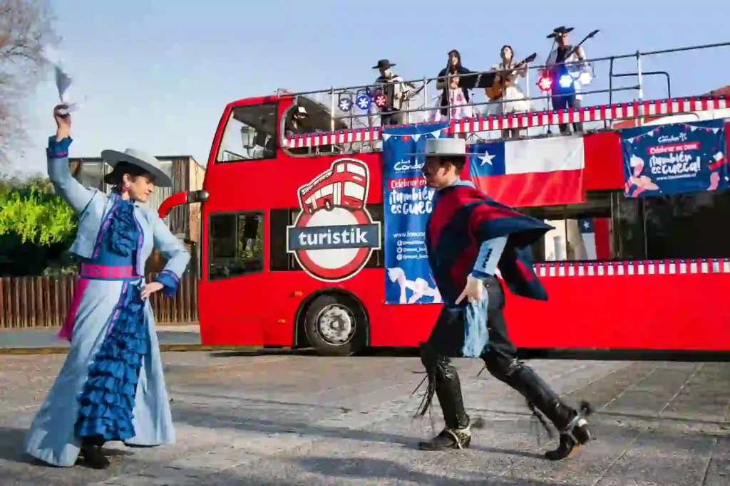 Pareja de bailarines con vestimenta tradicional chilena bailando cueca frente a un autobús rojo de TEM Turistik con músicos en vivo.