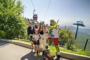 Familia posando junto a un personaje navideño disfrazado de Grinch en una atracción de TEM Turistik con teleféricos al fondo.